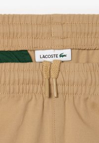 Bežīši ar elastīgu jostasvietu, ar Lacoste etiķeti un zaļu akcentu. Ietver bežīgas saites ar zeltītām galiem. Strukturēta auduma.
