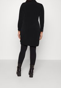 Abito nero con maniche lunghe e colletto, abbinato a leggings neri e stivali con tacco chunky neri. Tessuto liscio, design semplice.