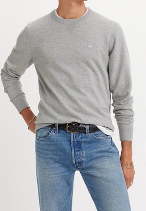 Man wearing light grey long-sleeve sweater, blue jeans with black braided belt, silver chain bracelet, and a tattoo on his right wrist.