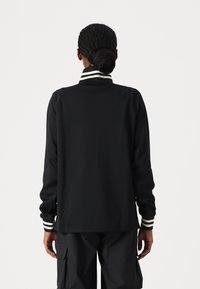 Crna jakna dugih rukava s visokim ovratnikom, sa bijelim prugastim detaljima na manžetama. Izrađena od glatkog materijala, dizajnirana za opušteni kroj.