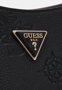 Materiale sintetico nero testurizzato con un logo triangolare in oro e nero con la scritta "GUESS USA" e un punto interrogativo. Embossing dettagliato visibile.