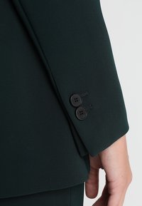 Rękaw ciemnozielonego żakietu o gładkim, teksturowanym wykończeniu, z dwoma czarnymi guzikami i subtelnym przeszyciem w okolicy mankietu.