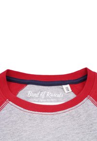Harmaa t-paita punaisilla raglan-hihnoilla ja tummansinisen kaula-aukon kanssa. Sisältää ommeltuja saumoja ja etiketin, jossa on "Band of Rascals" -brändäys.