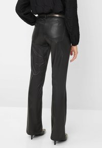 Pantalons évasés en cuir noir avec une texture lisse, dotés de poches arrière et d'une taille haute. Portés avec un blouse noire texturée.