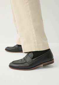 Zwarte leren loafers met een gestructureerde afwerking, voorzien van een gestikte accent en een lage hak, gedragen met crèmekleurige broek en zwarte sokken.