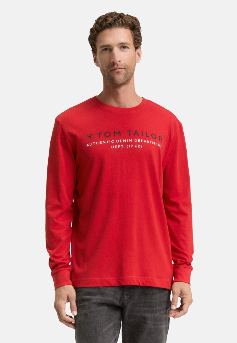 Mann mit lockigem Haar, der ein rotes Langarmshirt mit dem Aufdruck "Tom Tailor Authentic Denim Department" trägt, kombiniert mit dunklen Jeans vor einem weißen Hintergrund.