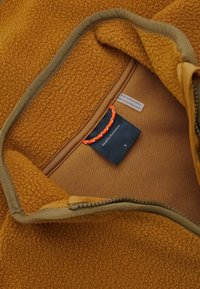 Fleecejacke mit braunem Außenmaterial, beigem Innenfutter und grünem Besatz. Verfügt über einen Reißverschluss und ein Etikett mit der Aufschrift "Peak Performance".