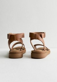Sandali in pelle marrone con un design minimalista, caratterizzati da fasce larghe, chiusura con fibbia sul retro e una soletta imbottita su una suola piatta.