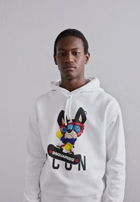 Weißer Hoodie mit einem bunten Cartoon-Hund-Design, der eine Brille und eine Mütze trägt, und dem Text "DSQUARED2" darunter. Baumwollmaterial, normale Passform.