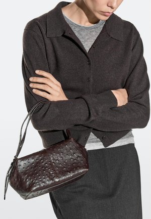 Person iført mørk cardigan og grå skjorte, der holder en lille tekstureret mørkebrun lædertaske med kort strop.