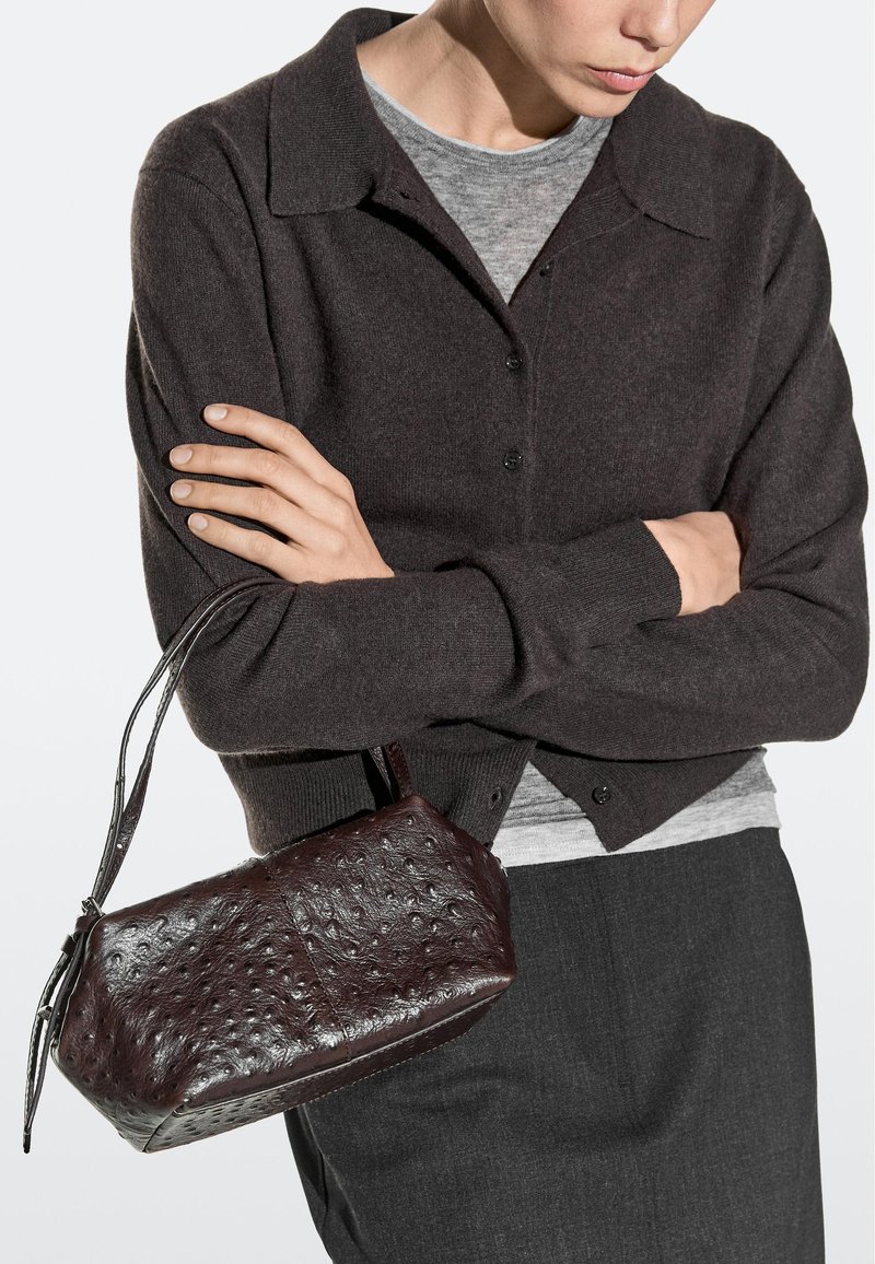 Personne portant un cardigan sombre et une chemise grise, tenant un petit sac à main en cuir brun foncé texturé avec une courte bandoulière.