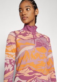 Long-sleeve zip-neck top in textured fabric featuring a mountain and forest pattern in shades of pink, orange, and beige.
