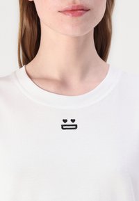Bílý tričko s kulatým výstřihem, které má černý vyšitý design ve stylu emoji: dvě srdce a usmívající se ústa umístěná na hrudi.
