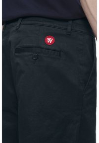 Black pants featuring a red circular logo patch, side and back pockets with a button closure, made from textured cotton fabric.
