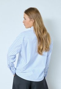 Langärmliges Hemd mit blau-weißen Streifen, Kragen, lockerem Schnitt und Rückenteil-Detail, kombiniert mit dunklen, weit geschnittenen Hosen.