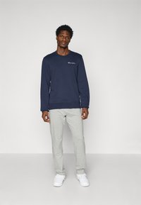 Marineblå sweatshirt med logo på brystet, parret med lysegrå sweatpants. Begge er lavet af blødt stof og har en afslappet pasform. Hvide sneakers.