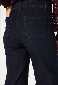 Jeans in denim blu scuro con vita alta, taglio dritto e due tasche posteriori. Il tessuto ha una superficie liscia e dettagli di cuciture sottili.