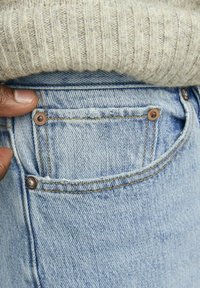 Jeans de denim azul claro con un bolsillo trasero visible, remaches de cobre y costuras en contraste. La tela presenta una textura de denim típica.