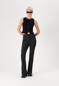 Haut noir court sans manches avec des boutons, associé à un pantalon évasé à rayures noires et blanches. Le modèle porte des lunettes de soleil sombres et des chaussures plates noires.