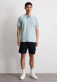 Helles blaues Baumwoll-Polohemd, marineblaue Cargo-Shorts, weiße Sneakers, steht vor einem neutralen Hintergrund. Verfügt über einen gerippten Kragen und kurze Ärmel.