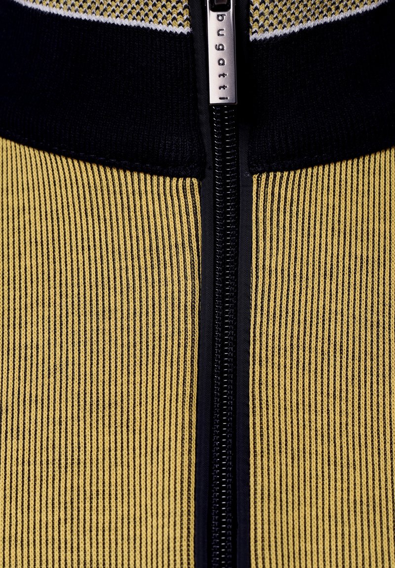 Giacca con zip in tessuto a righe verticali nera e gialla; presenta un'etichetta logo argento vicino alla zip e bordi scuri a contrasto.