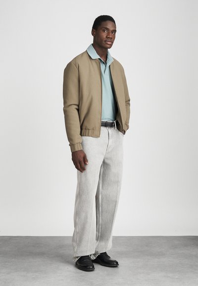 Beige bomberjakke over en lyseblå polo, kombinert med lysegrå baggy jeans og svarte sko. Enkelt design, uformell passform.