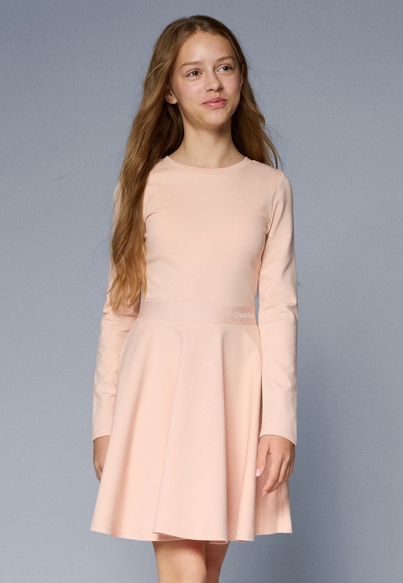 Vestido de manga longa, rosa claro, com corpete ajustado e saia ampla. Apresenta um logo da Calvin Klein na faixa da cintura. Tecido suave e leve.