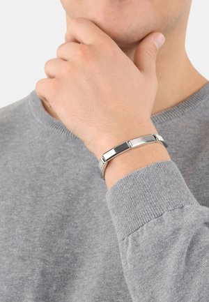 Braccialetto in metallo argento con maglie rettangolari, caratterizzato da superfici lisce e lucide. Indossato su una mano vicino a un maglione grigio.