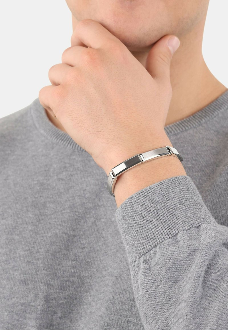Pulsera de metal plateado con eslabones rectangulares, que presenta superficies lisas y pulidas. Lleva en una mano cerca de un suéter gris.