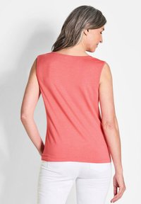 Ärmelloses korallenfarbenes Top aus weichem Stoff, mit einem runden Ausschnitt und geradem Schnitt. Getragen mit weißen Hosen, die das Rückendesign zeigen.