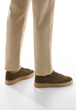 Sneakers in suede verde oliva con lacci marroni, punta arrotondata e suole in gomma color sabbia, abbinati a pantaloni beige chiaro su misura.