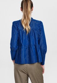 Blusa blu a maniche lunghe con strisce verticali, dettaglio arricciato sul retro, collo rotondo e polsini con bottone. Indossata sopra pantaloni marrone chiaro a fantasia.