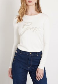 Maglione bianco a maniche lunghe con logo "Guess" in rilievo in strass; abbinato a jeans a vita alta blu scuro con bottoni dorati.