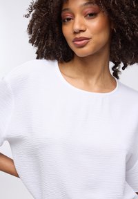 Top bianco texturizzato con scollatura rotonda e maniche corte, caratterizzato da un motivo ondulato su un tessuto leggero. Nessun accento visibile.