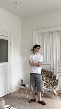 Balta trumpa rankovėmis marškinėliai, pilkos džinsinės šortai, rudos bateliai. Šviesus medinis grindys, baltos durys, raštuota kailinė antklodė ir dekoratyvūs daiktai.