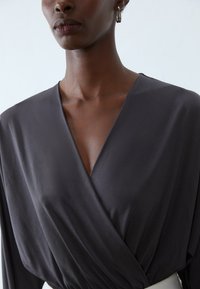 Mørk grå omslagsbluse med dyp V-hals, lange ermer og glatt tekstur. Minimalistisk design med subtile draperingsdetaljer.