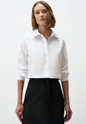 Chemise blanche à boutons avec col, manches longues retroussées aux poignets, associée à un pantalon noir ajusté. Tissu lisse avec une coupe classique.