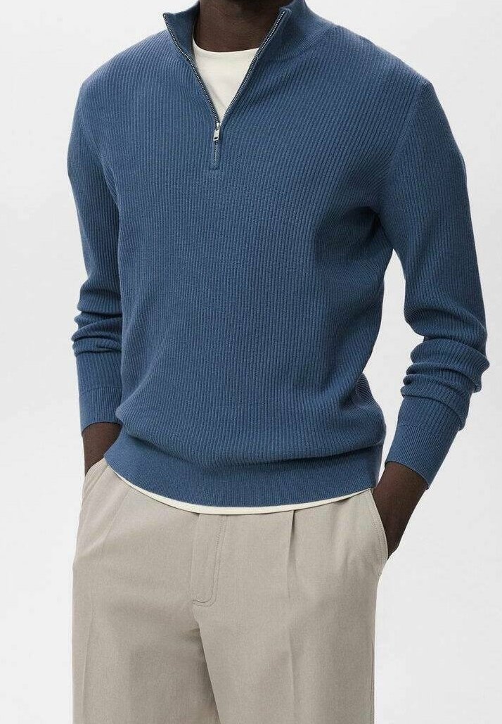 Homme portant un pull côtelé bleu à demi-fermeture éclair sur une chemise blanche, avec un pantalon beige plissé et les mains dans les poches.