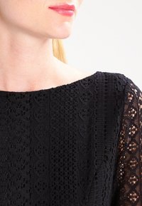 Sort blonderetop med detaljerede mønstre, tekstureret stof og en bred bådudskæring, samt gennemsigtige blonderærmer.