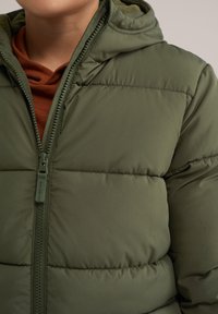 Zelena pufasta jakna z zadrgo, s kapuco in prešito teksturo. Nosi se nad oblačilom rjave barve spodaj.