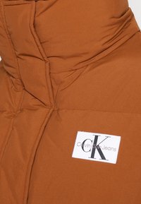 Doudoune marron avec col montant et patch logo rectangulaire blanc Calvin Klein Jeans sur le devant.