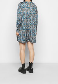 Šaty s květinovým vzorem v odstínech modré, hnědé a bílé, s dlouhými rukávy a klíčovou dírkou na zádech. Stylizováno s černými kotníkovými botami.