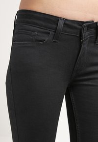 Jeans de mezclilla negra con un corte ajustado, que cuentan con un bolsillo frontal, cierre de botón metálico y cinturilla plana. Textura suave con detalles de costura mínimos.
