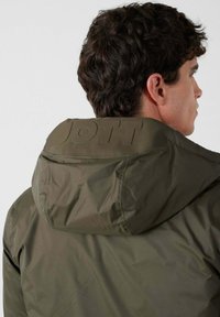 Giacca verde oliva con cappuccio, caratterizzata da un logo testurizzato sulla parte posteriore del collo. Il materiale sembra liscio e leggero.