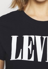 Černé tričko vyrobené z bavlny, s velkými bílými nápisy "LEVI". Krátké rukávy a kulatý výstřih zdůrazňují design.