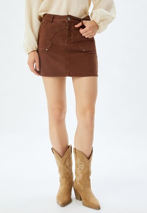 Gonna di denim marrone con tasche anteriori e passanti per cintura. Abbinata a stivali da cowboy beige con dettagli testurizzati e tacco grosso.