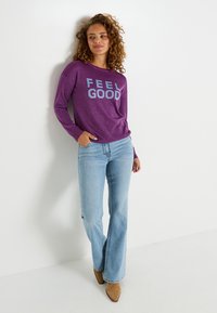 Chemise à manches longues pailletée violette avec le texte "FEEL GOOD" en bleu clair, associée à un jean bleu clair évasé et des bottes à talons beiges.
