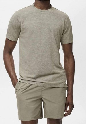 Homme portant un t-shirt à manches courtes marron clair et un short assorti, debout avec une main dans la poche sur un fond uni.