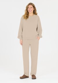 Beiger Sweatshirt mit gerippten Bündchen und Saum, kombiniert mit passenden, locker sitzenden Hosen. Das Modell trägt braune Clogs und steht vor einem schlichten Hintergrund.