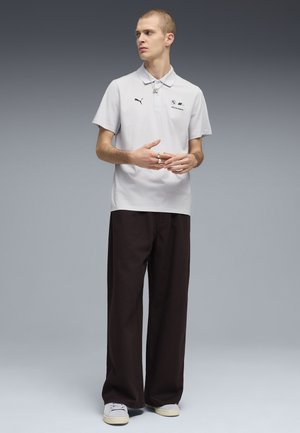 Młody mężczyzna w jasnoszarej koszulce polo BMW Motorsport, czarnych szerokich spodniach i białych tenisówkach, z srebrnymi pierścionkami, stoi na tle szarego gradientu.